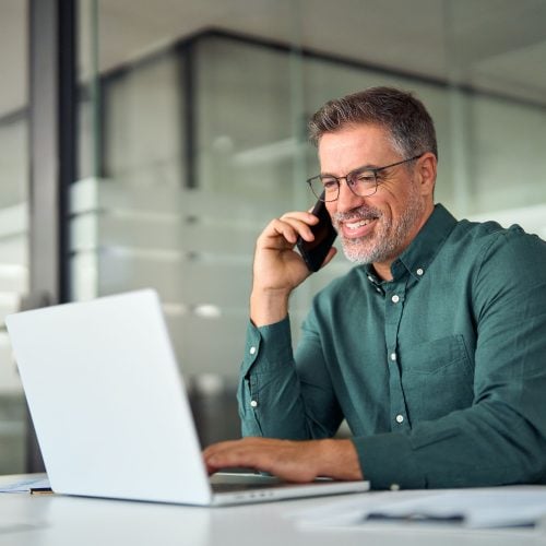 A smiling man with glasses sitting at a desk while talking on a smartphone and using a laptop.
