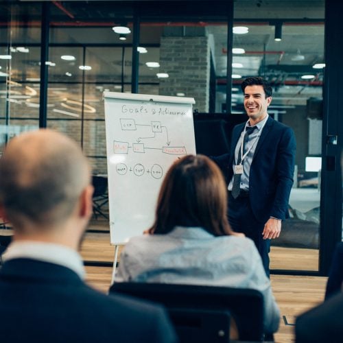 A smiling man in a suit presenting a "Goals & Achievements" flow chart on a whiteboard to a seated audience in a modern office.